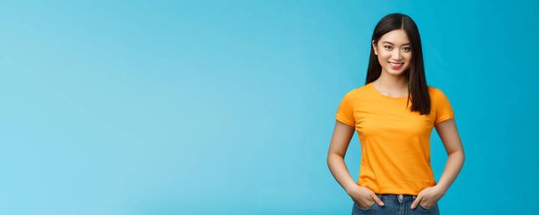 Selbstbewusst selbstbewusst lächelnd asiatische Frau stehen blauen Hintergrund Blick Kamera, halten die Hände Jeanstaschen Ausdruck selbstbewusster Stimmung, genießen positive Ergebnisse Hautpflege-Verfahren, tragen gelbes T-Shirt. - Foto, Bild