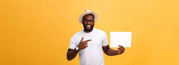 Picture of young smiling african-american man holding white blank board and pointing on it, on yellow background, copy space. - Photo, Image