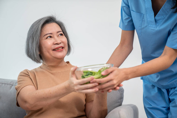A female nurse serves a bowl of salad to a contented senior couple. Health care and medical assistance for the elderly, nursing home for pensioners. - Photo, image