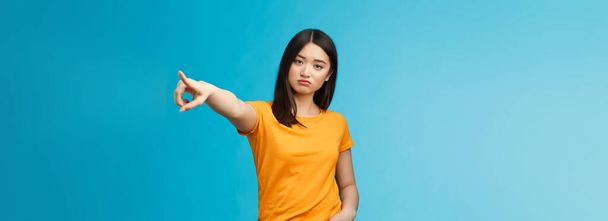 Go away from here. Serious-looking arrogant displeased bossy asian girl showing way-out, pointing sideways index finger look camera demand leave, stand blue background ignorant. Copy space - Photo, Image