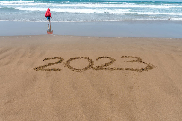 Year 2023 written in the sand on the beach with a boy in a red coat and the sea in the background. - Zdjęcie, obraz
