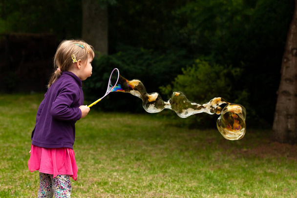 Feliz niño en edad escolar alegre soplando enormes burbujas de jabón al aire libre, joven chica caucásica haciendo una gran burbuja de jabón afuera en el parque, solo. Divertirse, actividades de verano gente estilo de vida - Foto, Imagen