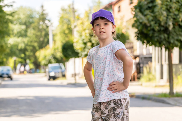 Young confident school age child, girl in a baseball cap standing on the street outdoors, hands on sides, kid confident posture, portrait, copy space, blurred background, one person. People lifestyle - Photo, Image
