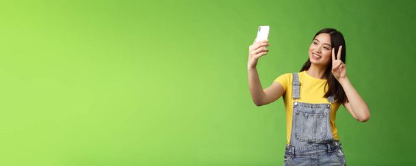 Tender schattig aziatische brunette vrouw, hold smartphone, poseren selfie, kijken telefoon scherm, fotograferen, tonen overwinning vrede teken, vrolijk chatten vriend in het buitenland, staan groene achtergrond. Kopieerruimte - Foto, afbeelding