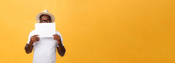 Young happy African-american hiding behind a blank paper, isolated on yellow background. - Foto, imagen