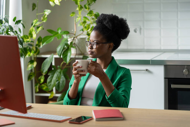 Επιτυχημένη σκεπτόμενη Αφρο-Αμερικανίδα που πίνει καφέ κάνοντας διάλειμμα κατά τη διάρκεια της δουλειάς και σκέφτεται τρόπους επίλυσης επιχειρηματικών προβλημάτων. Κορίτσι με κομψό κοστούμι κάθεται στο τραπέζι με τον υπολογιστή στο γραφείο στο σπίτι - Φωτογραφία, εικόνα