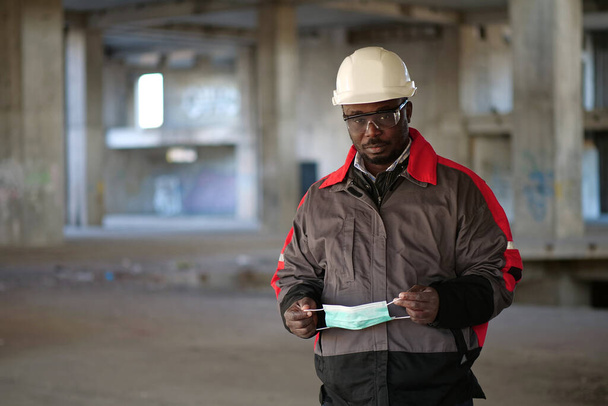 Africano americano construtor fica no local de construção, olha para a câmera e segura em mãos máscara protetora médica - Foto, Imagem