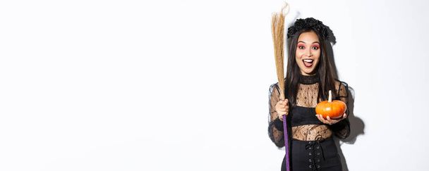 Podekscytowana atrakcyjna azjatycka dziewczyna świętuje Halloween, nosi strój czarownicy, trzyma dynię i miotłę, idzie cukierek lub psikus, stoi na białym tle. - Zdjęcie, obraz