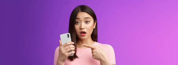 Preocupado confuso bonito jovem asiático mulher segurar smartphone boca aberta, apontando telefone dedo, olhar questionado chateado, perturbado ver estranho post online, ficar fundo roxo. Espaço de cópia - Foto, Imagem