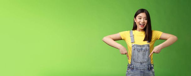 Живая энергичная азиатская милая женщина продвигает рекламу указывая пальцами вниз, показать вам прохладное место, улыбающаяся камера радостно, рассказывая большие приятные новости стенд зеленый фон эмоциональный. - Фото, изображение