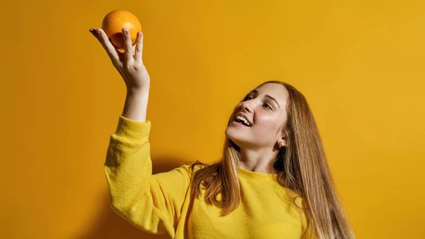 Cropped image of european female teenage holding and looking at whole orange in hand. Girl of zoomer generation. Healthy eating. Modern youngster lifestyle. Yellow background. Studio shoot. Copy space - Foto, afbeelding