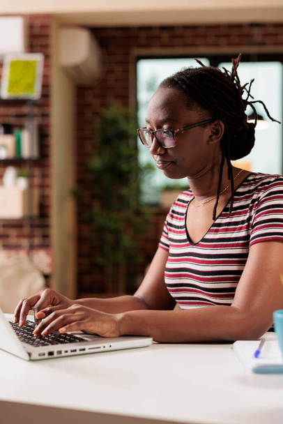 Masasında oturan Afrikalı Amerikalı kadın bir dizüstü bilgisayar üzerinde çalışıyor. Üniversitedeki Webinar kursunda internetten öğreniyorum. Bilgisayar ekranına yoğunlaşmış şekilde bakılıyor. Bilgi arıyor, araştırıyor. - Fotoğraf, Görsel