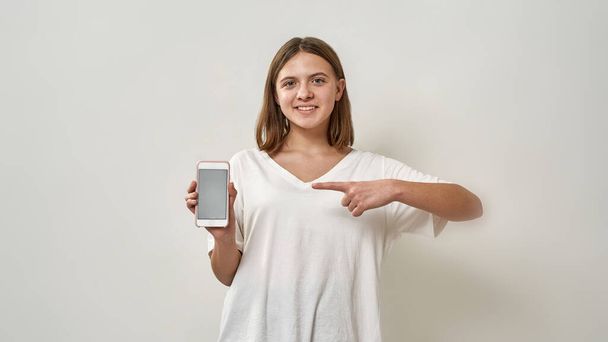 Front view of smiling european teenage girl showing smartphone and looking at camera. Blonde female of zoomer generation. Modern youngster lifestyle. Isolated on white background in studio. Copy space - Photo, image