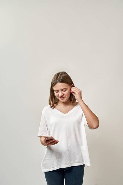 Smiling caucasian teenage girl put earphone in ear and choose music for listen in smartphone. Blonde female of zoomer generation. Modern youngster lifestyle. White background. Studio shoot. Copy space - Photo, Image