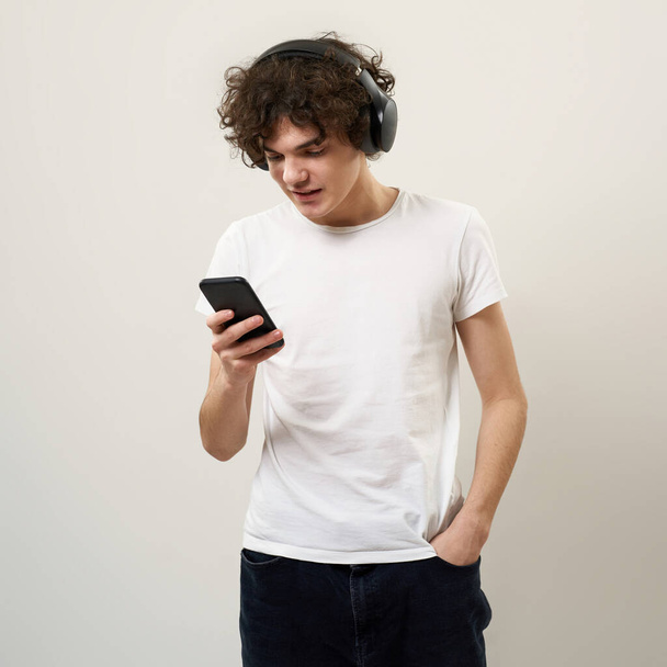 Caucasian teenage boy with hand in pocket using smartphone and listening music in headphones. Curly guy of zoomer generation. Modern youngster lifestyle. White background. Studio shoot. Copy space - Zdjęcie, obraz