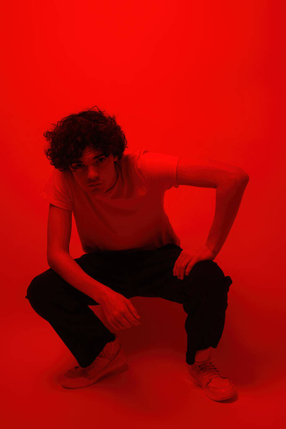 Confident caucasian teenage boy sitting and looking at camera. Curly guy of zoomer generation. Concept of modern youngster lifestyle. Isolated on red background. Studio shoot. Copy space - Fotografie, Obrázek