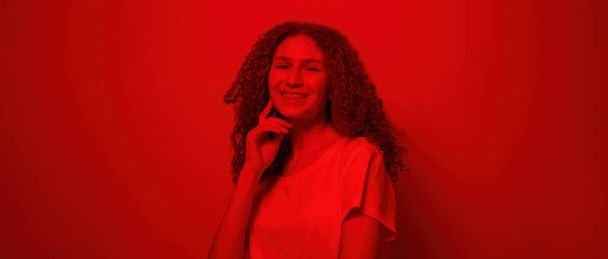 Portrait of smiling caucasian female teenage looking at camera. Curly girl of zoomer generation wearing t-shirt. Concept of modern youngster lifestyle. Isolated on red background in studio. Copy space - Foto, Imagem