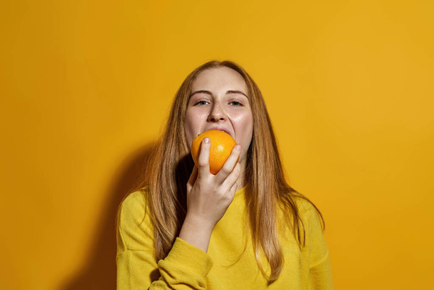 Partial image of caucasian female teenage eating orange and looking at camera. Girl of zoomer generation. Healthy eating. Concept of modern youngster lifestyle. Yellow background in studio. Copy space - Foto, afbeelding