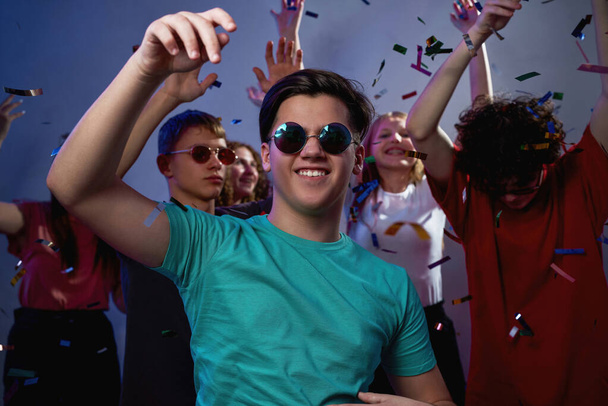 Boldog kaukázusi tinédzser srác szemüvegben táncol és szórakozik a barátaival repülő konfettiben lila háttérrel. Ifjúsági ünnep és buli. Szórakozás és kikapcsolódás. Éjszakai élet. Stúdió fotózás - Fotó, kép