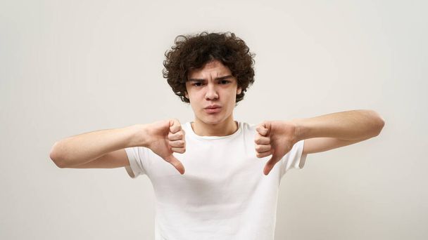 Dissatisfied caucasian teenage boy showing thumbs down and looking at camera. Curly guy of zoomer generation. Concept of modern youngster lifestyle. Isolated on white background in studio. Copy space - Foto, imagen