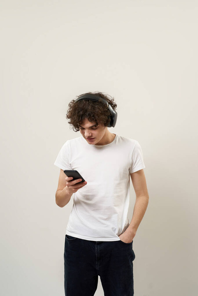 Caucasian teenage boy with hand in pocket choosing music on smartphone for listening in headphones. Curly guy of zoomer generation. Modern youngster lifestyle. White background in studio. Copy space - Photo, image