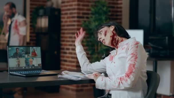 Scary looking office zombie waving hand at video teleconference on laptop. Spooky evil monster with bloody and deep scars greeting online conference members while sitting in office workspace. - Footage, Video
