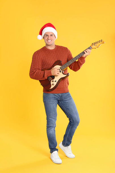 Man in Santa hat playing electric guitar on yellow background. Christmas music - Zdjęcie, obraz