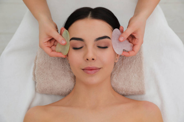 Young woman receiving facial massage with gua sha tools in beauty salon, top view - Fotó, kép
