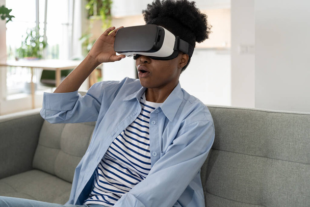 Shocked African American woman using vr glasses at home. Virtual reality and wearable tech concept with young black female wearing headset goggles. Digital technology for entertainment or leisure - Photo, Image