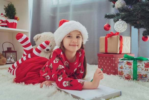Frohe Weihnachten und frohe Feiertage. Ein kleines fröhliches Mädchen in rotem Rock, gestreiften Socken, mit Nikolausmütze auf dem Boden unter dem Weihnachtsbaum mit Geschenken schreibt Briefe an den Weihnachtsmann. Blickt in die Kamera. - Foto, Bild