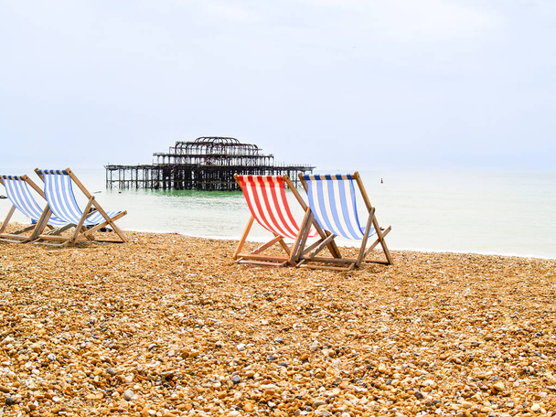 Brighton Beach és jellegzetes csíkos strand székek kék vagy piros és fehér.és maradványai régi Brighton Pier csak off-shore. - Fotó, kép