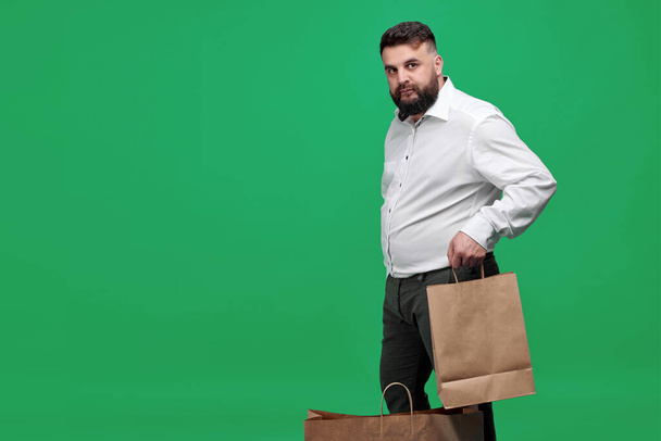 A Caucasian-looking man with a brown paper bag on a green background. A man with a white shirt with shopping bags. Studio shot of a man with purchases. - Fotografie, Obrázek
