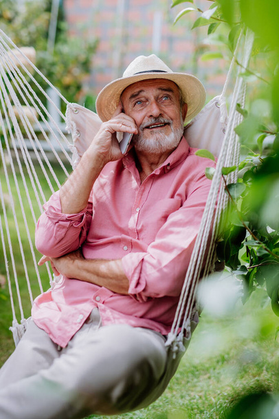 Χαρούμενος πρεσβύτερος κάθεται σε μια κούνια στον κήπο και phoneing. - Φωτογραφία, εικόνα