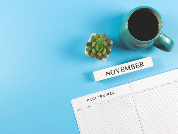 Bovenaanzicht of platte lay-of gewoonte tracker boek met houten kalender november, blauwe kop zwarte koffie en sappige plantenpot op blauwe achtergrond met kopieerruimte. - Foto, afbeelding