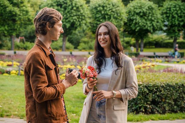Tiro de jovem casal em passeio com flores e fotocâmera em dia ensolarado no parque de verão. - Foto, Imagem