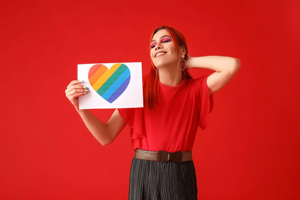 Stylish transgender woman holding paper sheet with rainbow heart against red background - Photo, Image