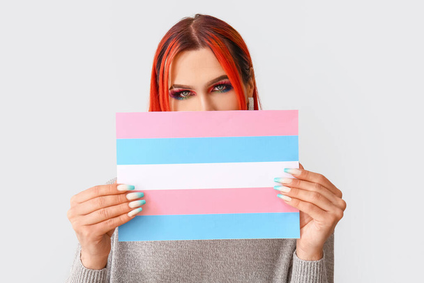 Elegante mujer transgénero sosteniendo bandera transgénero contra fondo claro - Foto, Imagen