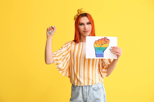 Stylish transgender woman holding paper sheet with clenched rainbow fist against yellow background - Photo, Image