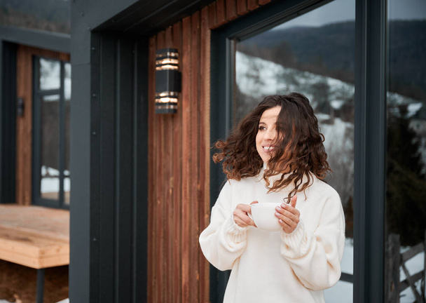 Curly young woman resting on terrace of modern barn house in the mountains. Happy female tourist holding cup of tea, enjoying in new cottage in winter. - Foto, afbeelding