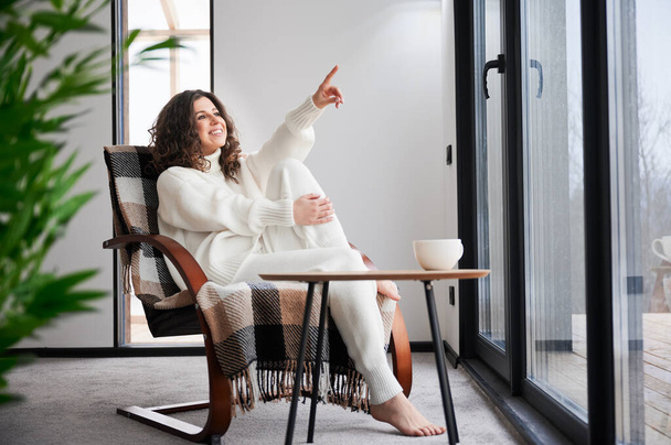 Curly young woman enjoying weekends inside contemporary barn house. Happy female tourist sitting on chair, pointing through panoramic windows in new cottage. - Foto, Bild
