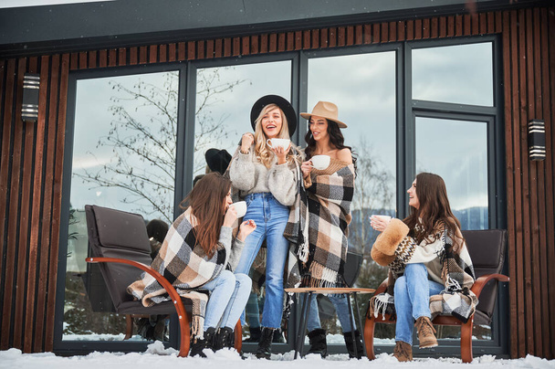 Young women enjoying winter weekends on terrace of contemporary barn house. Four girls having fun, sitting on chairs and drinking hot tea. - Photo, Image