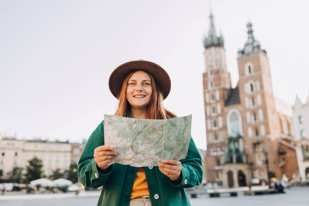 Ελκυστική νεαρή τουρίστρια εξερευνά νέα πόλη. Redhead κορίτσι κρατώντας ένα χάρτη στην πλατεία Αγοράς στην Κρακοβία. Ταξιδεύοντας στην Ευρώπη το φθινόπωρο. - Φωτογραφία, εικόνα
