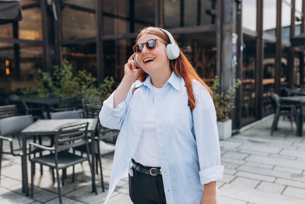 Прекрасная счастливая молодая женщина в солнечных очках слушает музыку через беспроводные наушники на городском фоне. Любитель музыки наслаждается музыкой. Девушка ходит по городской улице - Фото, изображение