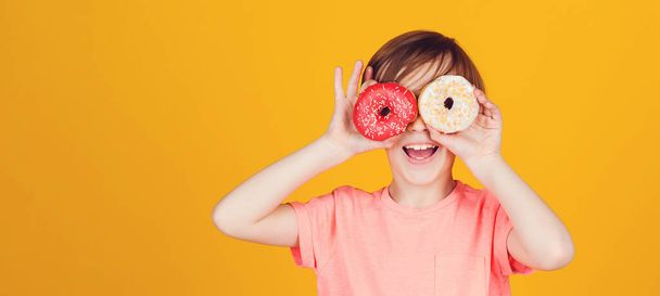 Happy child played with donuts on yellow background wall. Kid enjoying sweets. Cute funny boy holding donuts eyes. Birthday party. Tasty food for kids. Colored donuts. Fashion, people, lifestyle. - Photo, Image