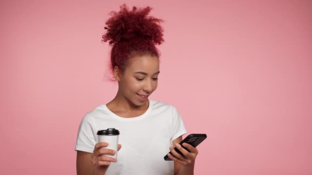 Közelkép mosolygós afro-amerikai üzletasszony gépelés böngészés okostelefon nézi a kamerát. Portré vörös hajú göndör nő dolgozik mobiltelefon online távoli izolált rózsaszín háttér - Felvétel, videó