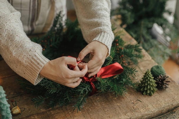 Žena ruce zdobí vánoční věnec s červenou stuhou na venkovském dřevěném stole s borovicovými kužely a svíčkami. Dělat vánoční věnec, náladový sváteční image. Prázdninový workshop, zimní výzdoba - Fotografie, Obrázek