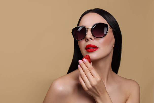 Attractive woman in fashionable sunglasses holding strawberry against beige background. Space for text - Фото, изображение