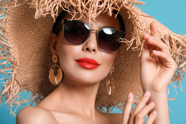 Attractive woman in fashionable sunglasses and wicker hat against light blue background, closeup - Foto, Bild