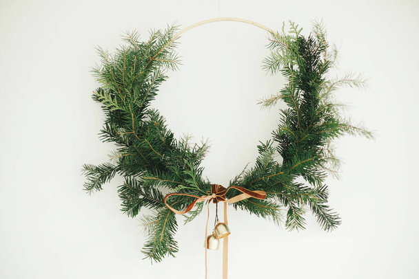 Merry Christmas and Happy Holidays! Modern Christmas wreath with velvet ribbon and golden bells hanging on white wall background. Winter holiday decor, moody image. Boho wreath - Photo, Image
