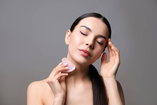 Beautiful young woman doing facial massage with gua sha tool on grey background - Fotó, kép